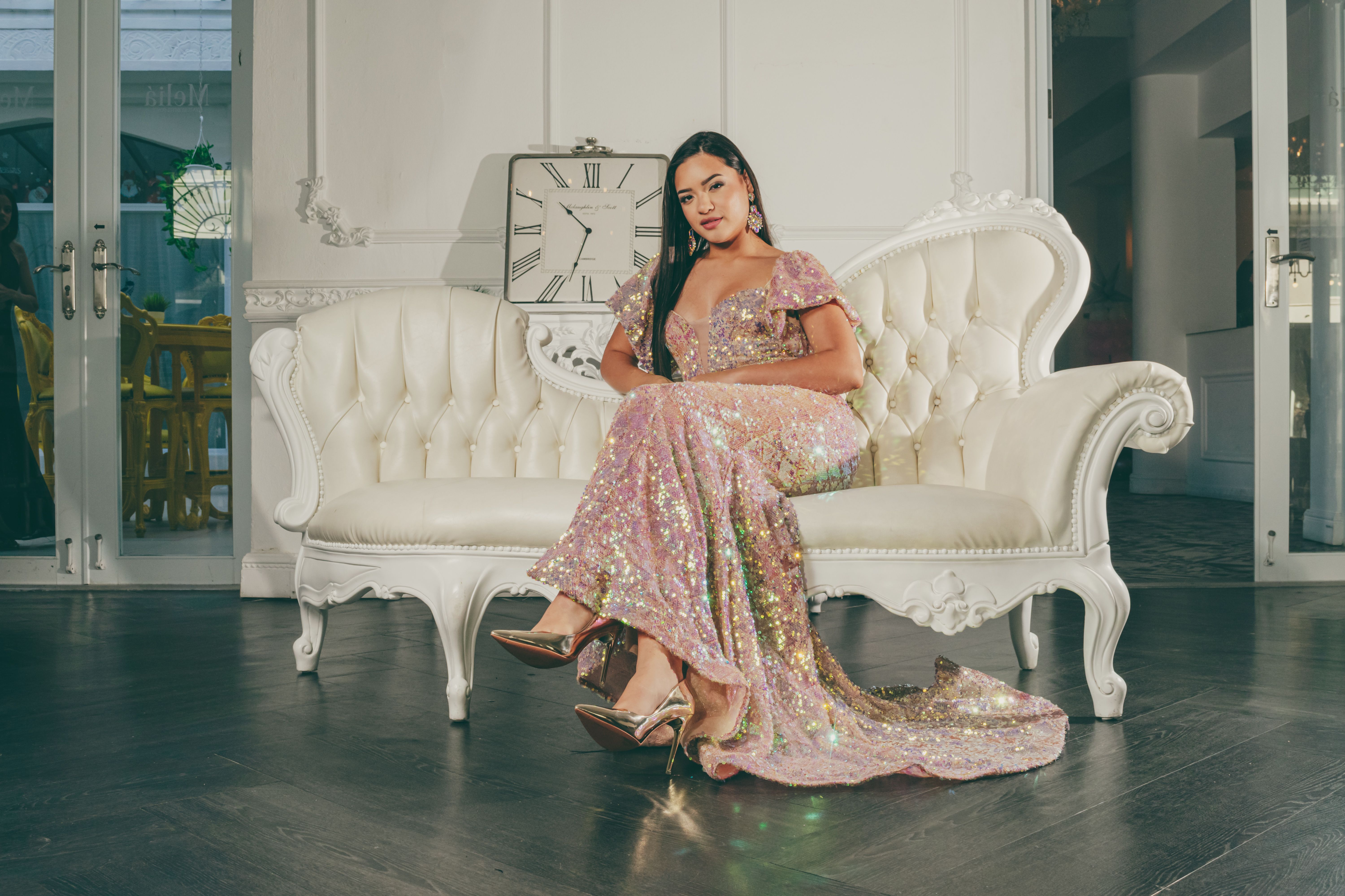 A woman in a glittery gown sits elegantly on a white sofa in a stylish indoor setting.