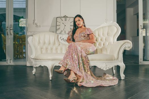 A woman in a glittery gown sits elegantly on a white sofa in a stylish indoor setting.
