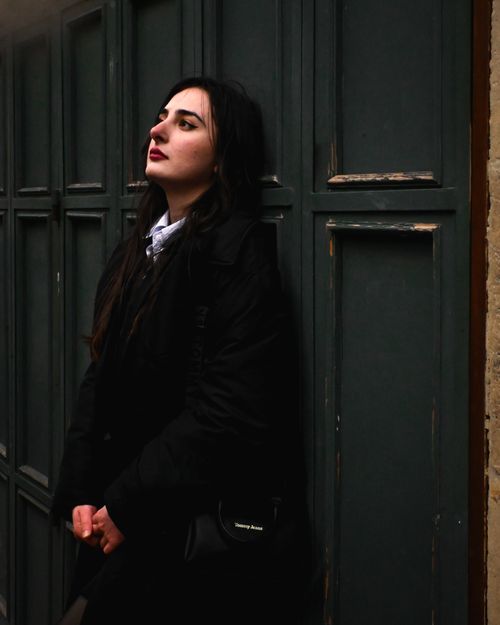 Free A woman in a black coat leans against a green wooden wall, looking contemplative. Stock Photo