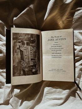 An open Jane Austen book on bed sheets, bathed in warm sunlight creating dramatic shadows.