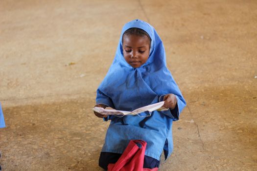 A young girl in a blue hijab sits on the floor reading a book, capturing a peaceful moment of learning.