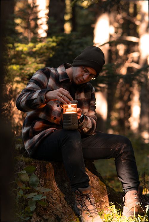 Free A man in a hat lights a lantern while sitting on a tree trunk in a forest setting. Stock Photo