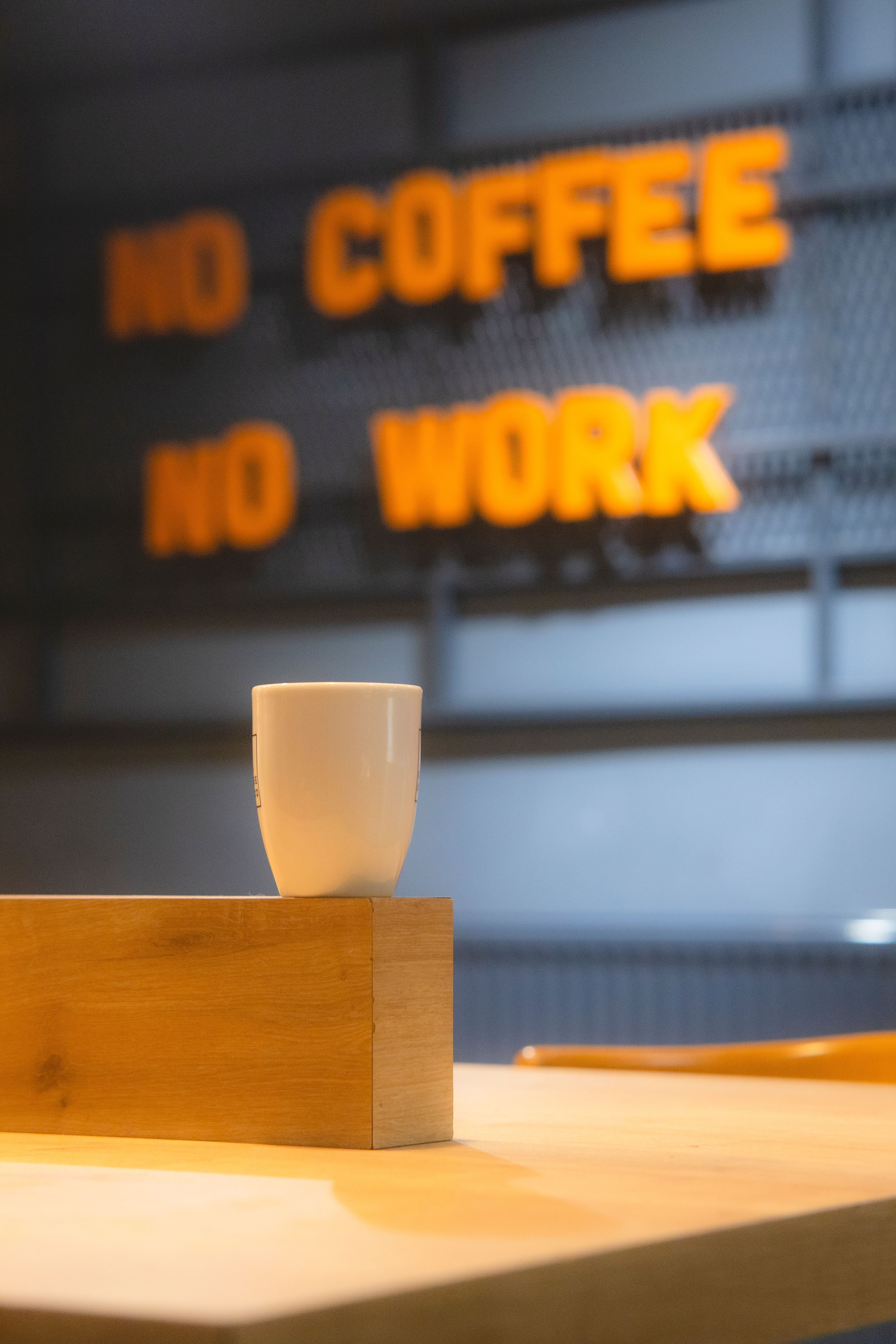 White cup on wooden surface with blurred motivational sign 'No Coffee No Work' in background.