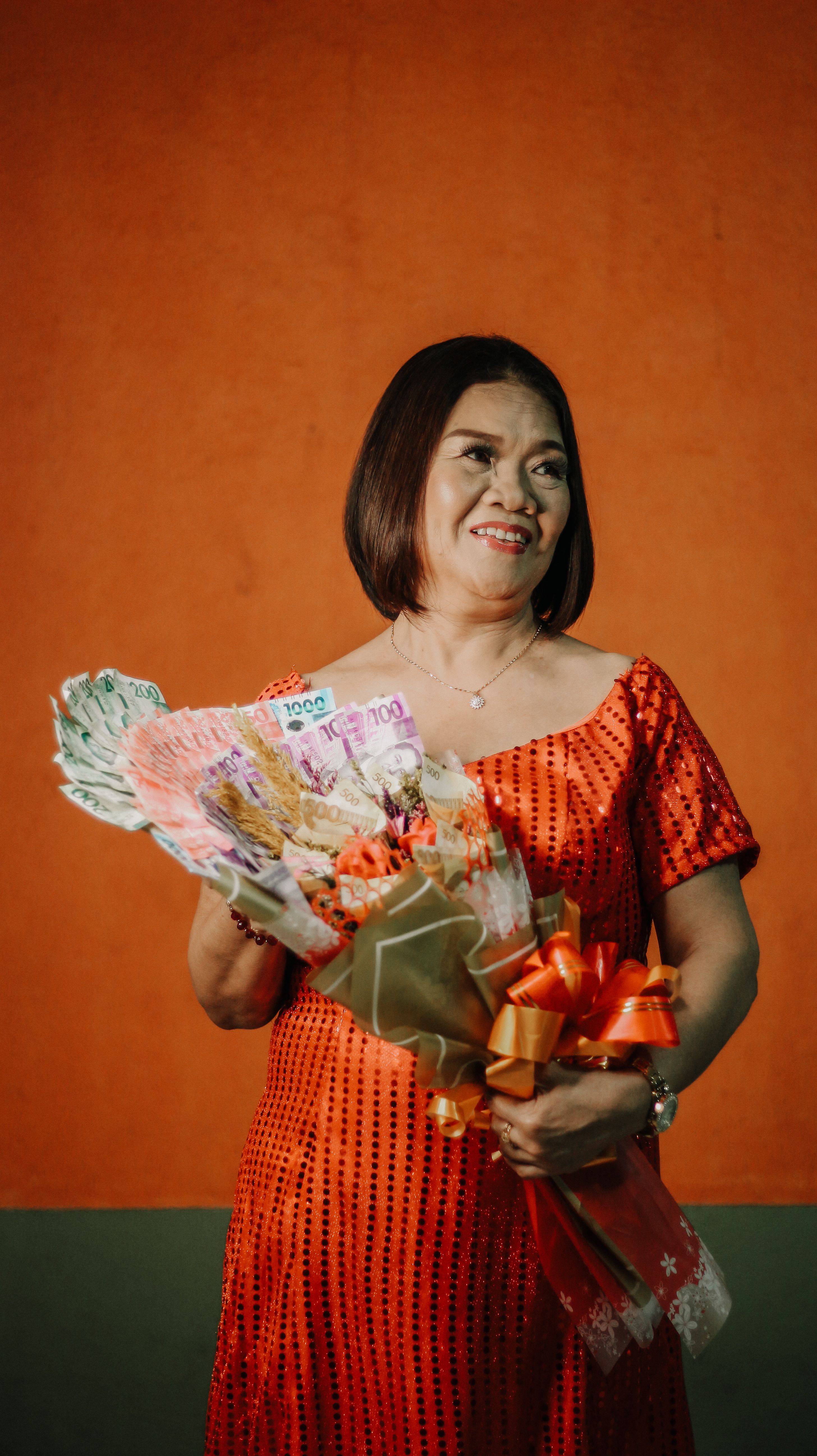 Stylish woman in vibrant orange dress holding a money bouquet against an orange backdrop.