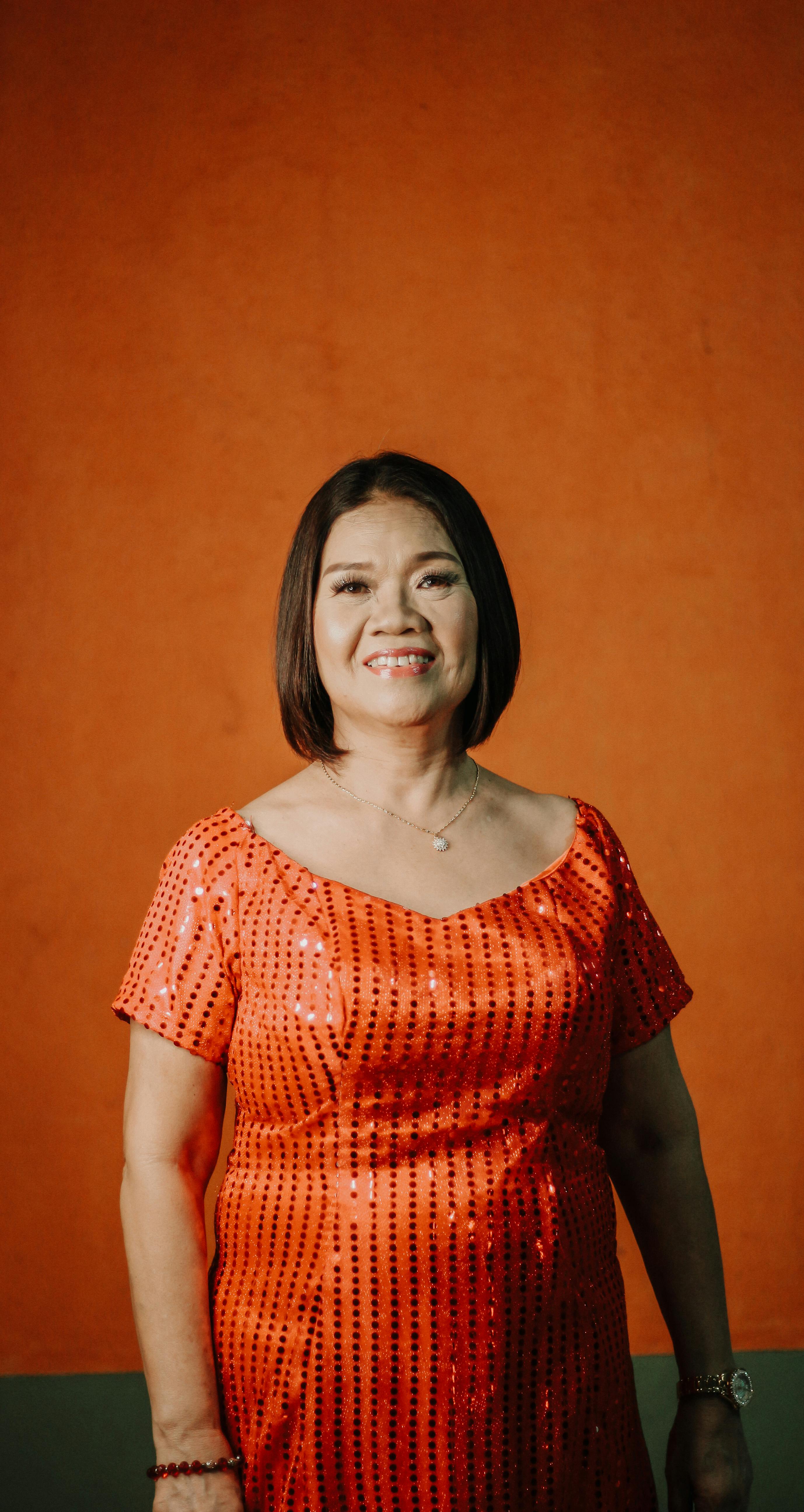 Smiling woman in an elegant orange dress against a simple backdrop, showcasing fashion and beauty.