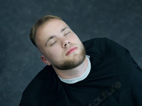 A serene portrait of a young man with a beard and closed eyes, wearing a black shirt.