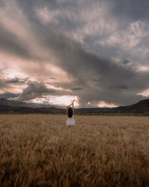 A woman stands with arms raised in a wheat field under a moody sunset sky, capturing serenity and freedom.