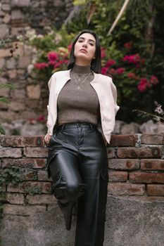 Fashionable woman in leather pants and jacket posing by brick wall with flowers.