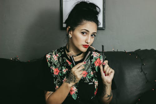 Woman in Floral Shirt Sits on Black Leather Sofa and Holds Make-up Brushes