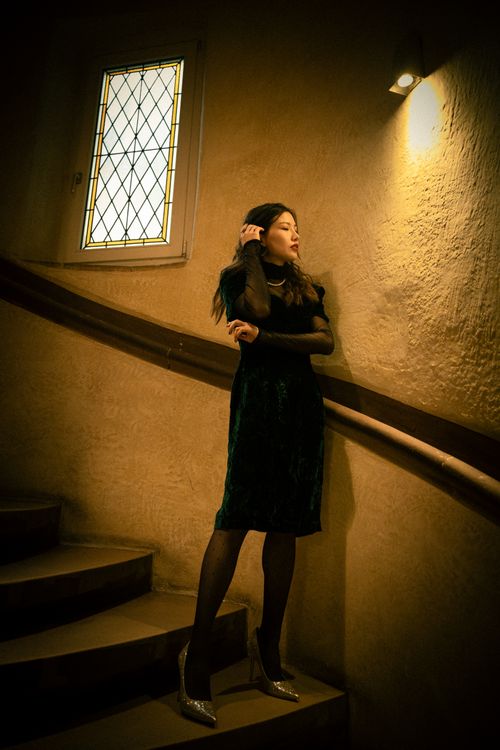 Free Chic woman in a green dress poses elegantly on indoor spiral stairs under warm lighting. Stock Photo