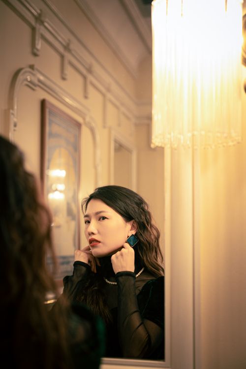 Free Stylish woman in black dress adjusting earrings, captured in mirror reflection with soft, warm lighting. Stock Photo