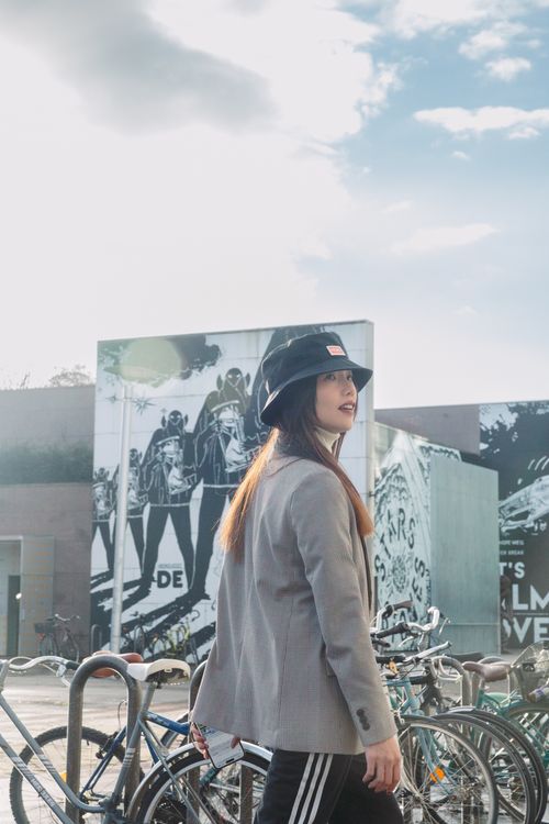 Free Young woman in a suit jacket and hat standing near bicycles in an urban setting. Stock Photo