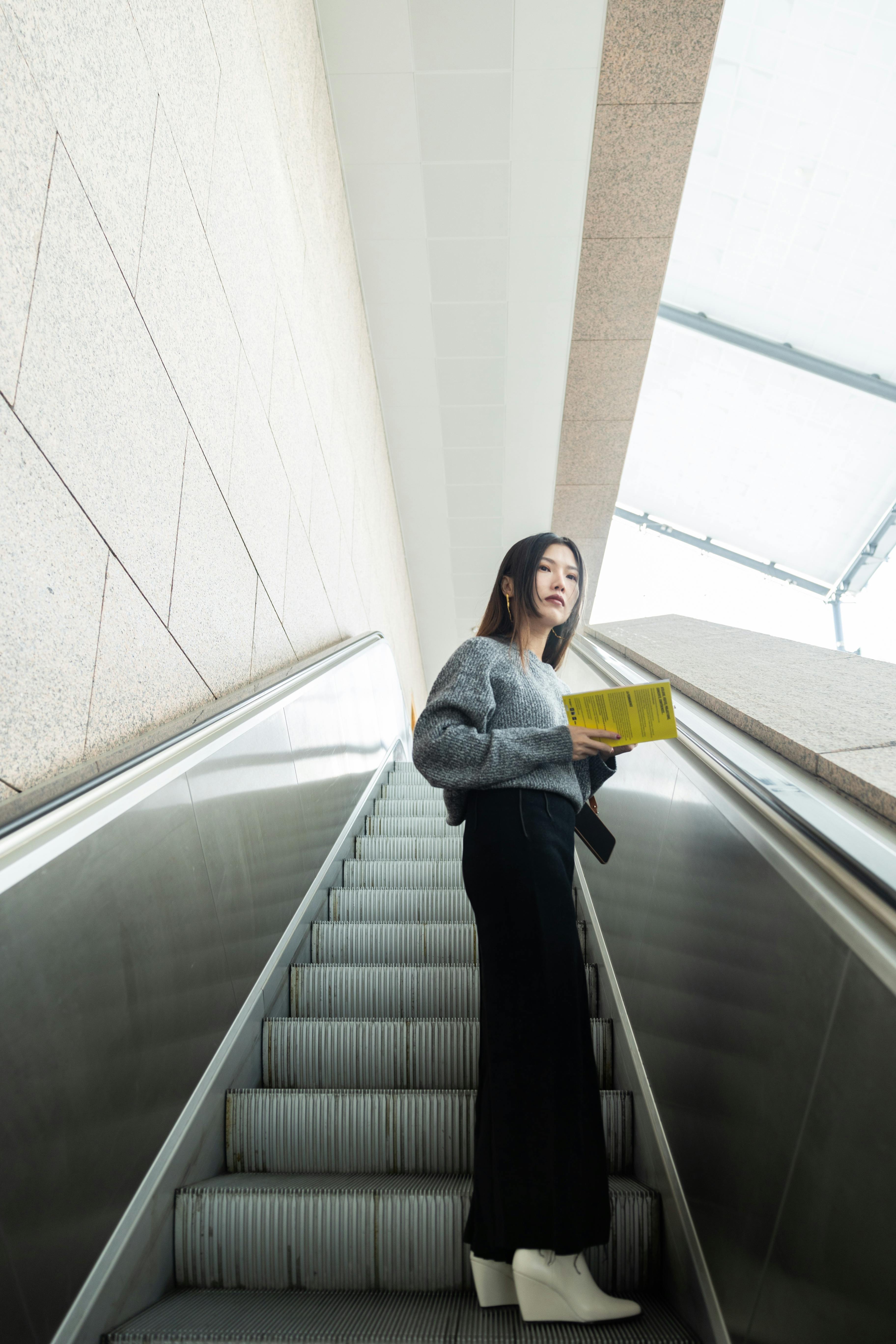 Woman dressed in modern attire holds a book while ascending an indoor escalator.