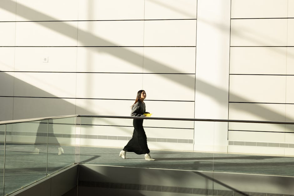 Willow Go vs Elvie Stride: Hands-Free Breast Pump Showdown A woman in stylish attire walks through a minimalist sunlit corridor, casting dynamic shadows.