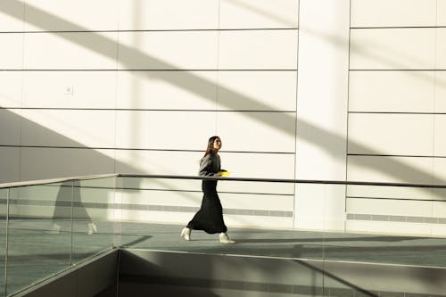 A woman in stylish attire walks through a minimalist sunlit corridor, casting dynamic shadows.