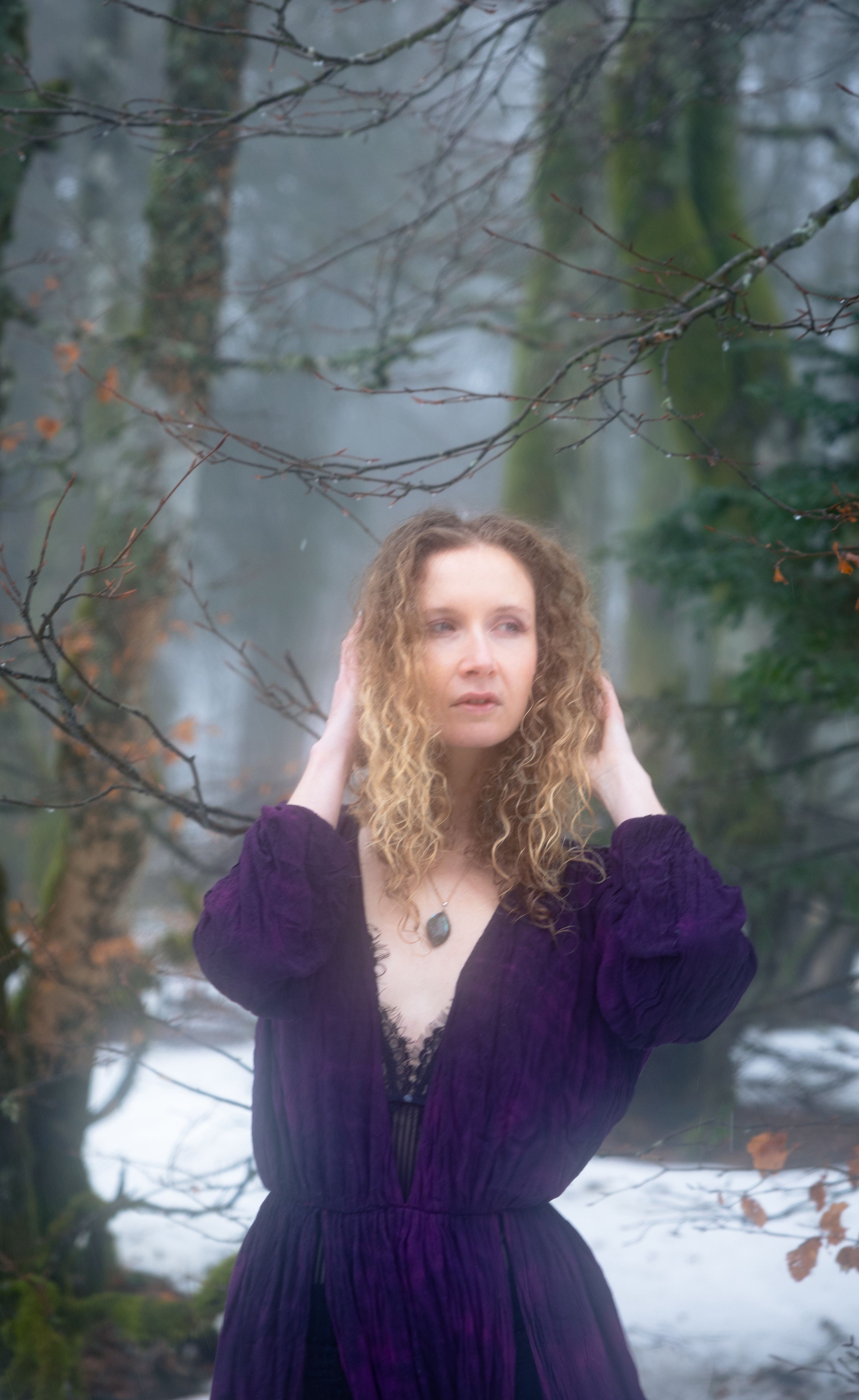 A woman in a purple dress poses in a foggy winter forest, exuding elegance and mystery.