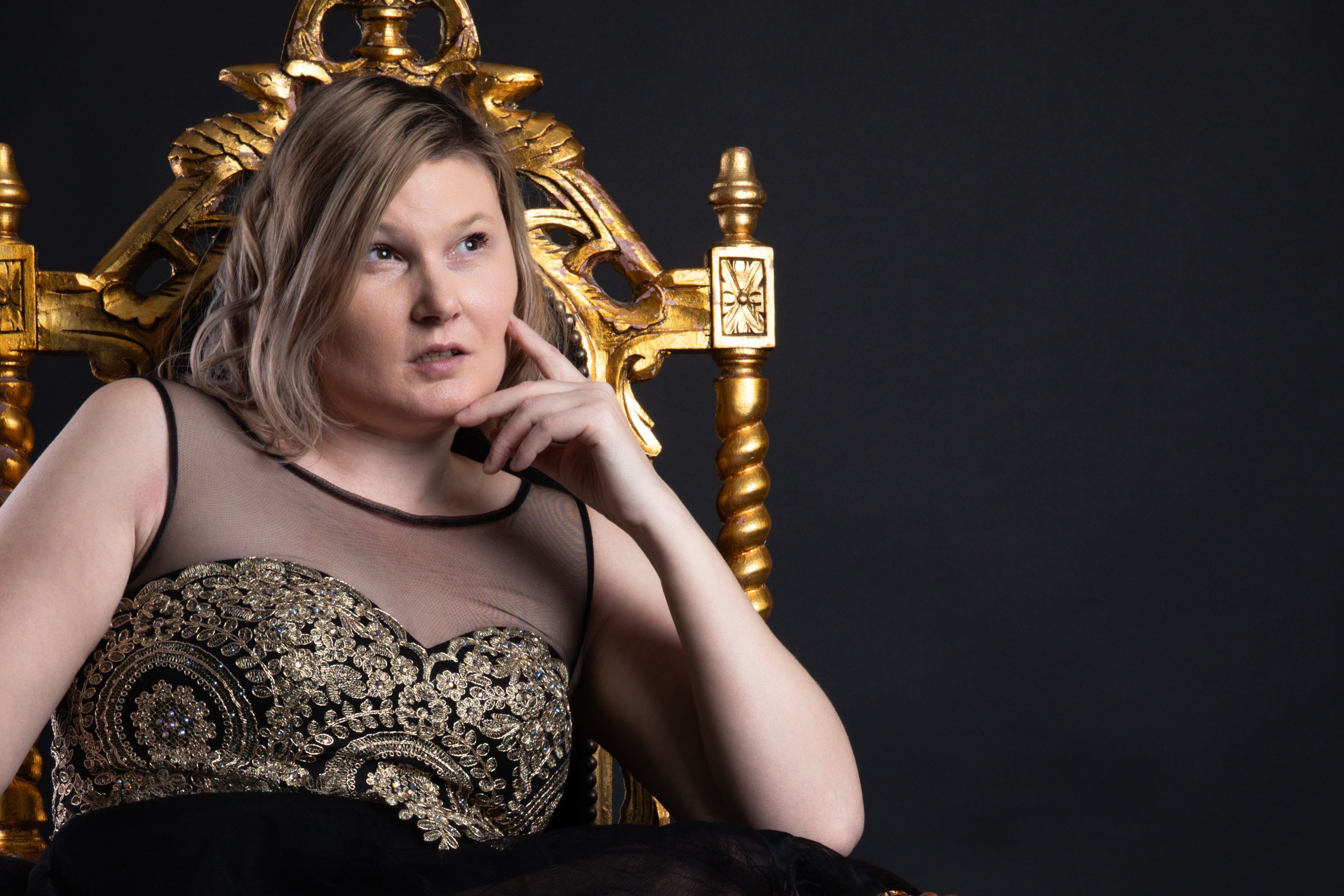Young woman in a black and gold dress sitting in an ornate chair.