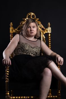 A young woman in a black and gold dress sits on an ornate chair against a dark background.
