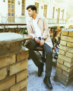 A fashionable man in a beige coat seated outdoors at a café patio, exuding urban style.