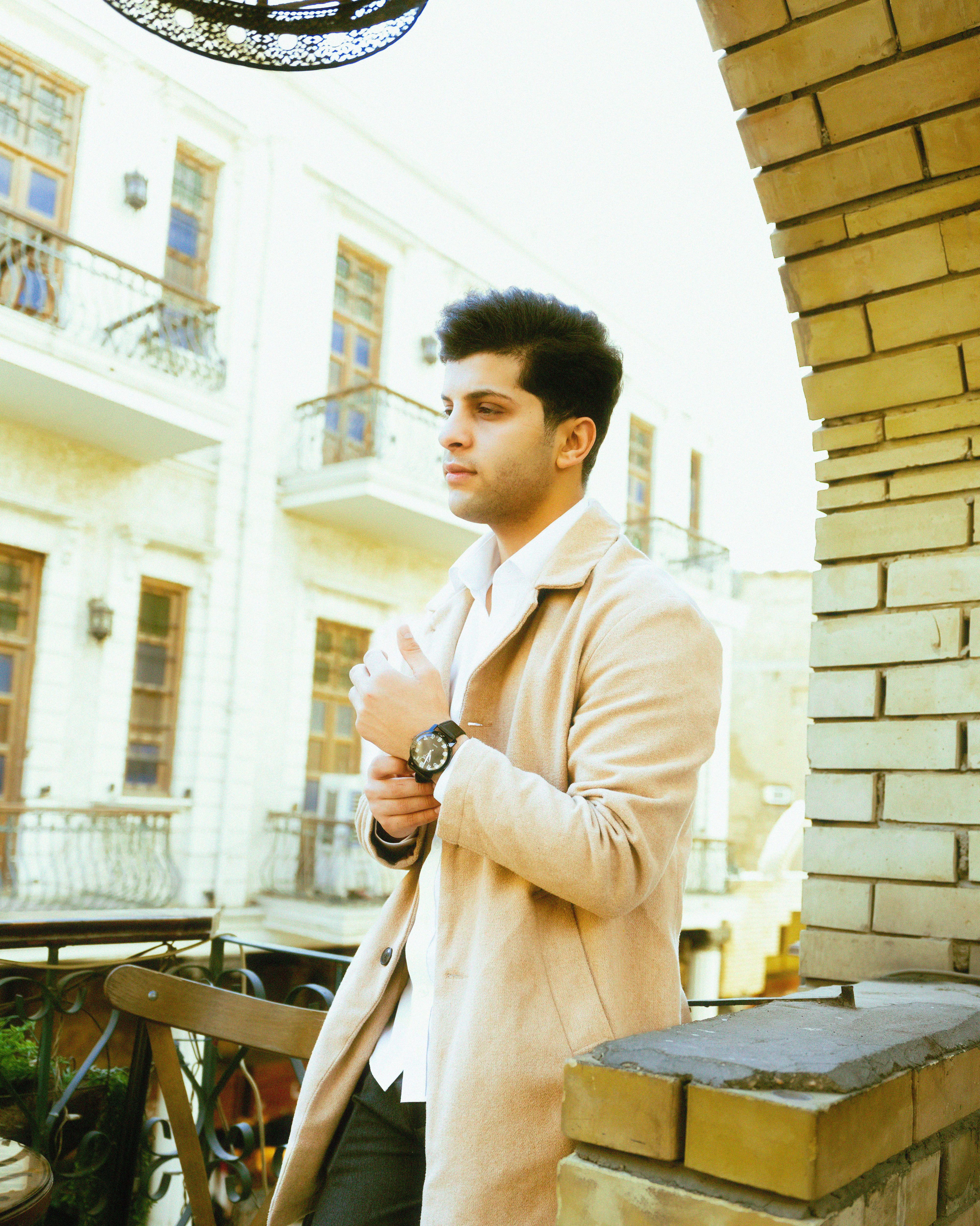Portrait of a young man in a coat enjoying autumn sunlight on a balcony with urban architecture in the background.