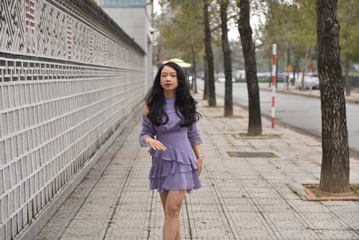 A fashionable woman strolls along a city sidewalk in a lilac dress, embodying urban style.