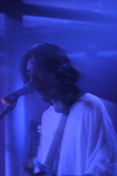 A shadowy guitarist sings into a microphone under blue stage lights, capturing the essence of live music.