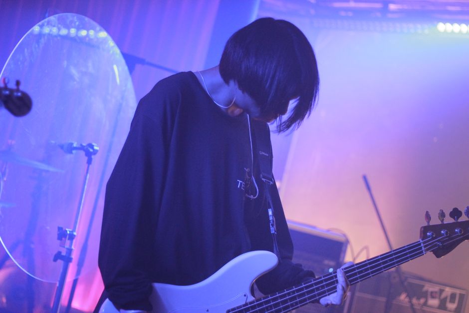 A musician plays an electric bass guitar on stage under purple lighting.