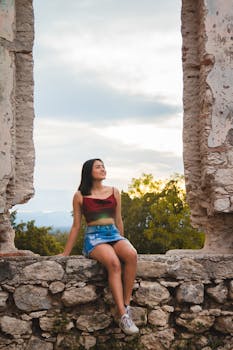 A young woman sits smiling on a stone ruin with a scenic background, enjoying the outdoors.