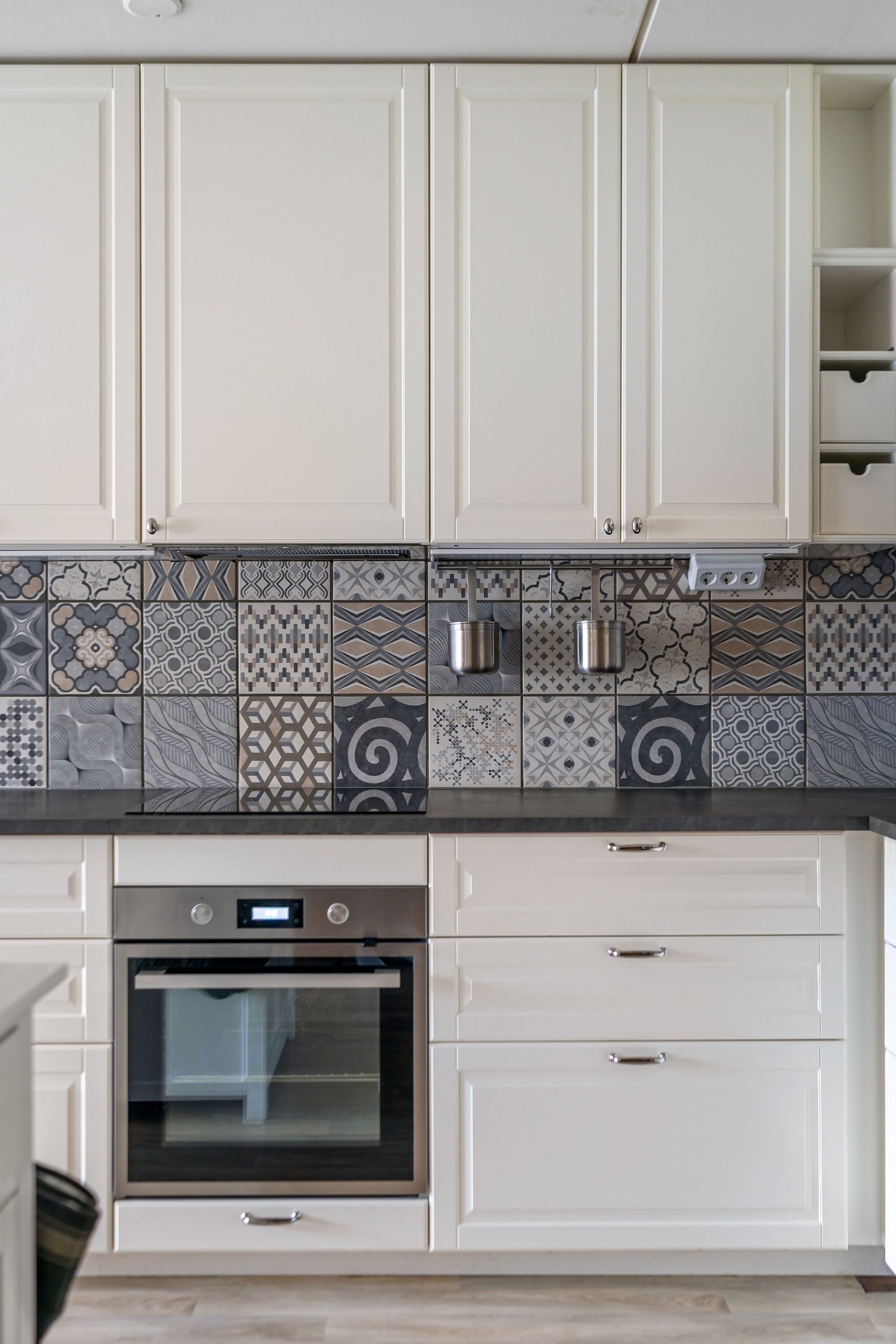A completed kitchen section with white raised-panel cabinetry, a patterned grey tiled splashback, dark benchtop, built-in oven and drawers.