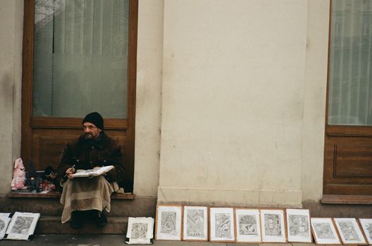A street artist in Budapest sits by the sidewalk with his artworks on display against a building wall.