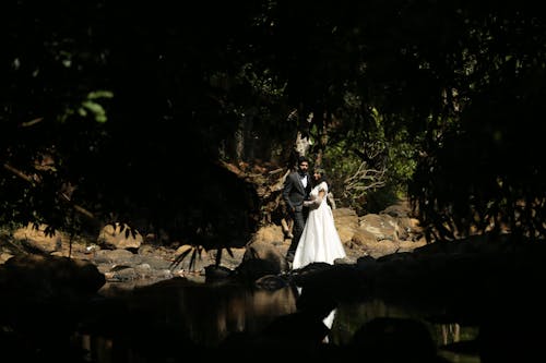 Newlyweds Standing under Trees and by Stream in Forest