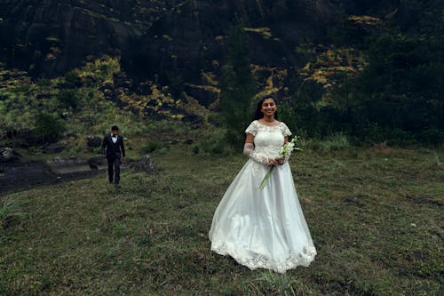 Smiling Woman in Wedding Dress Standing with Flowers and Man Walking behind