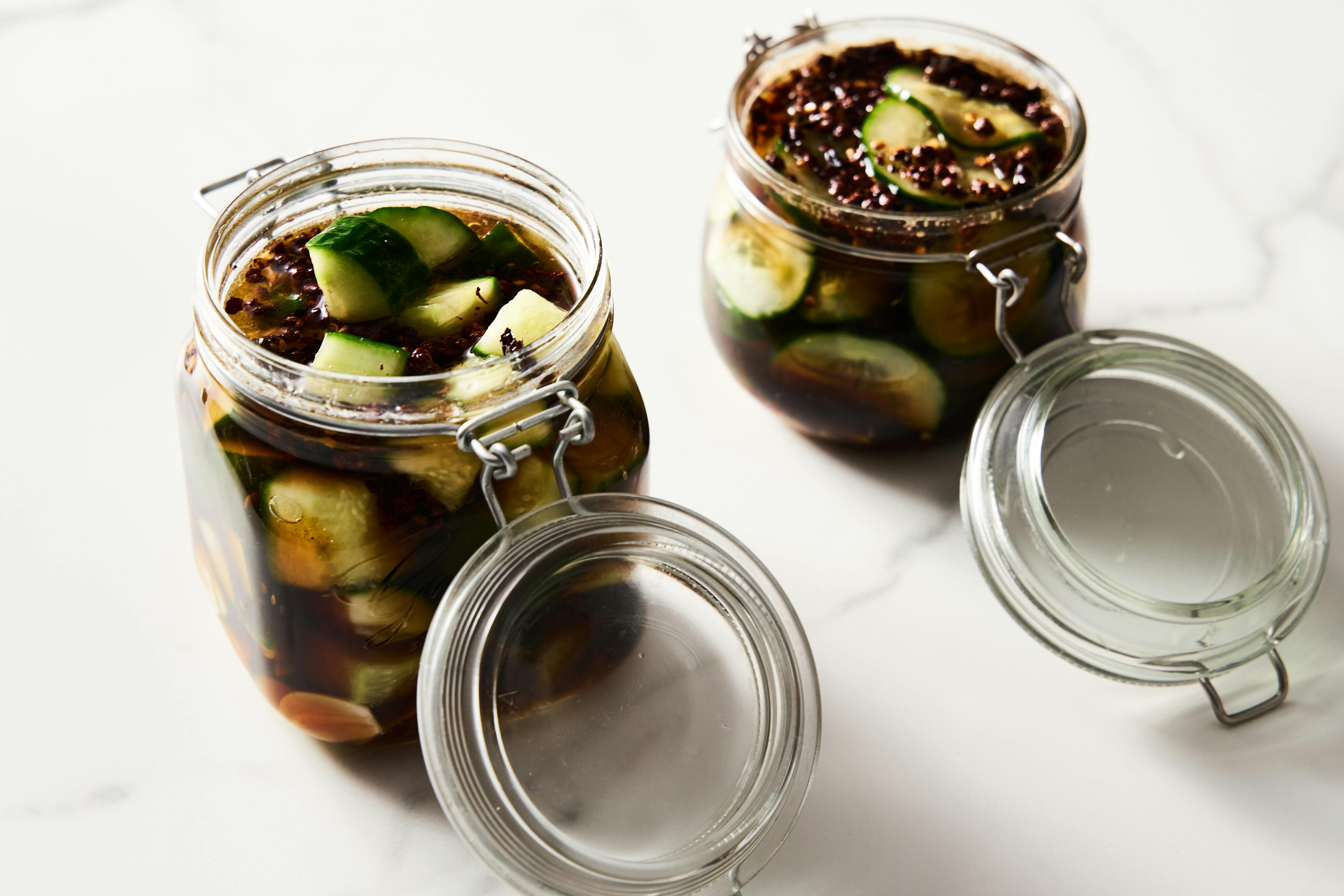 Two jars of sliced cucumbers pickled with spices and herbs on a white surface.