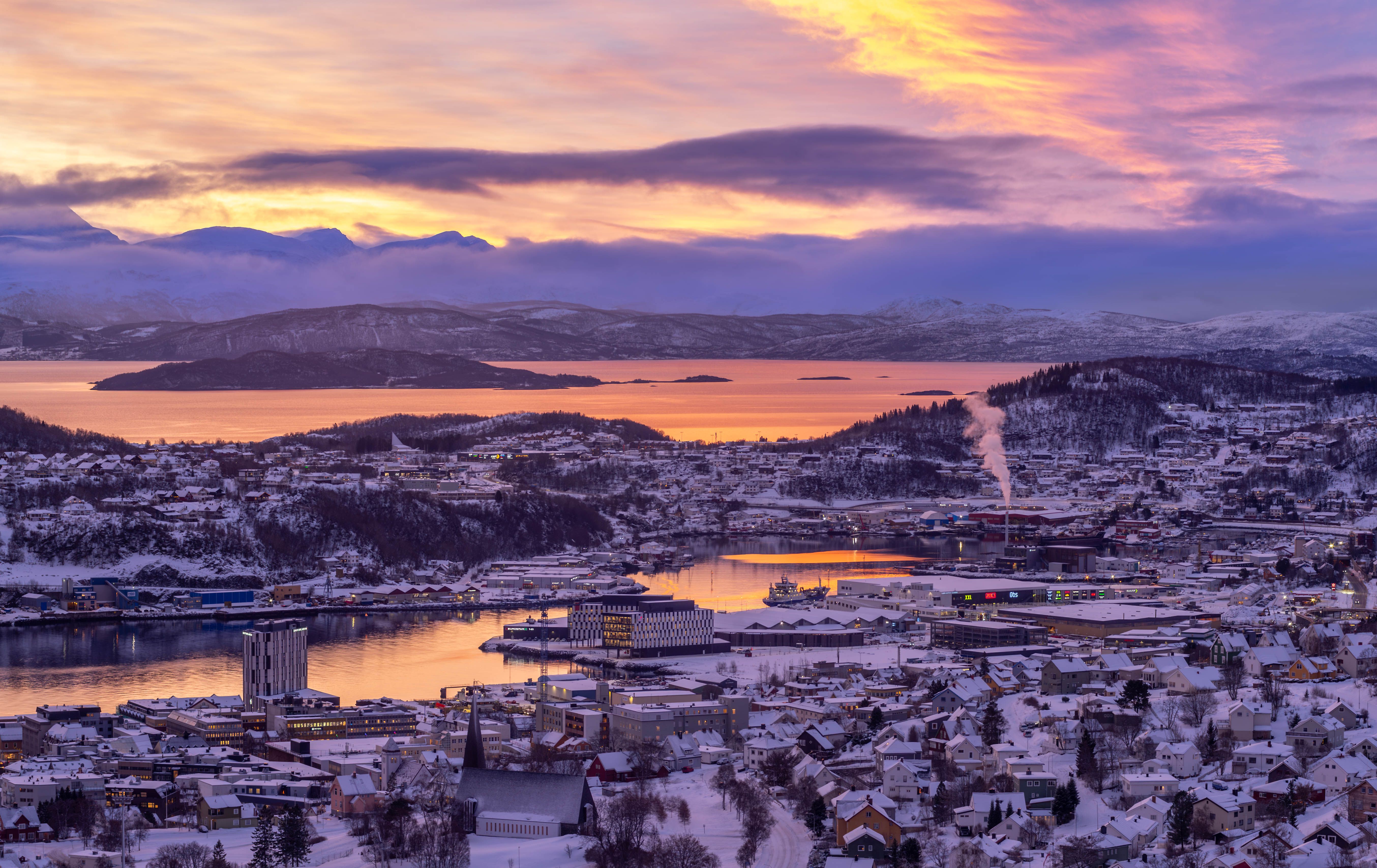 Experience a serene winter sunset over the snow-covered town of Harstad, with colorful skies reflecting on the tranquil waters.