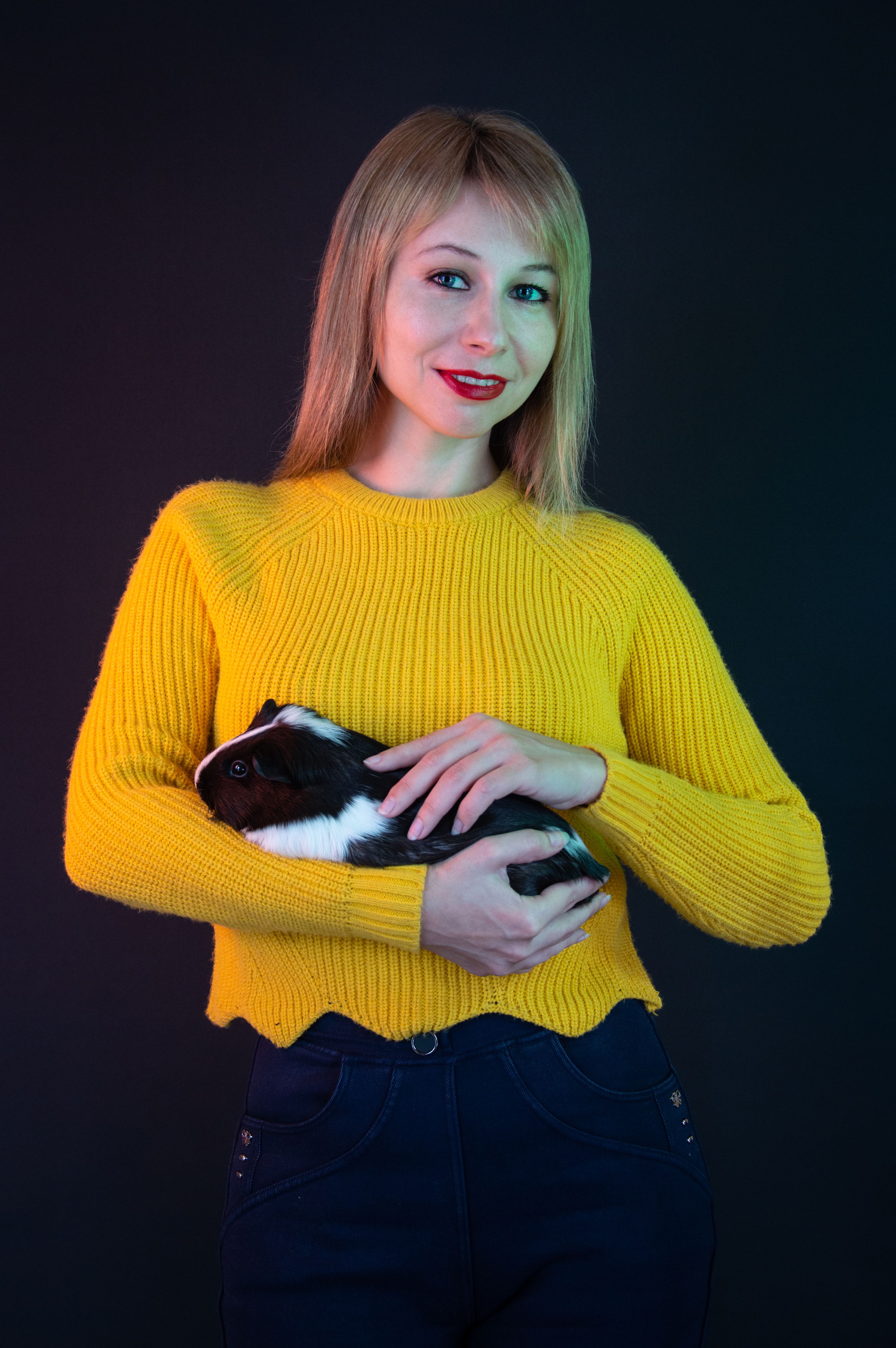 A smiling woman in a bold yellow sweater gently holding a guinea pig against a dark background.