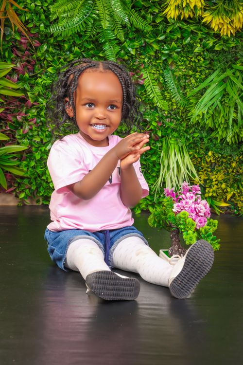 Free Adorable child smiling while sitting indoors with a lush floral backdrop. Stock Photo