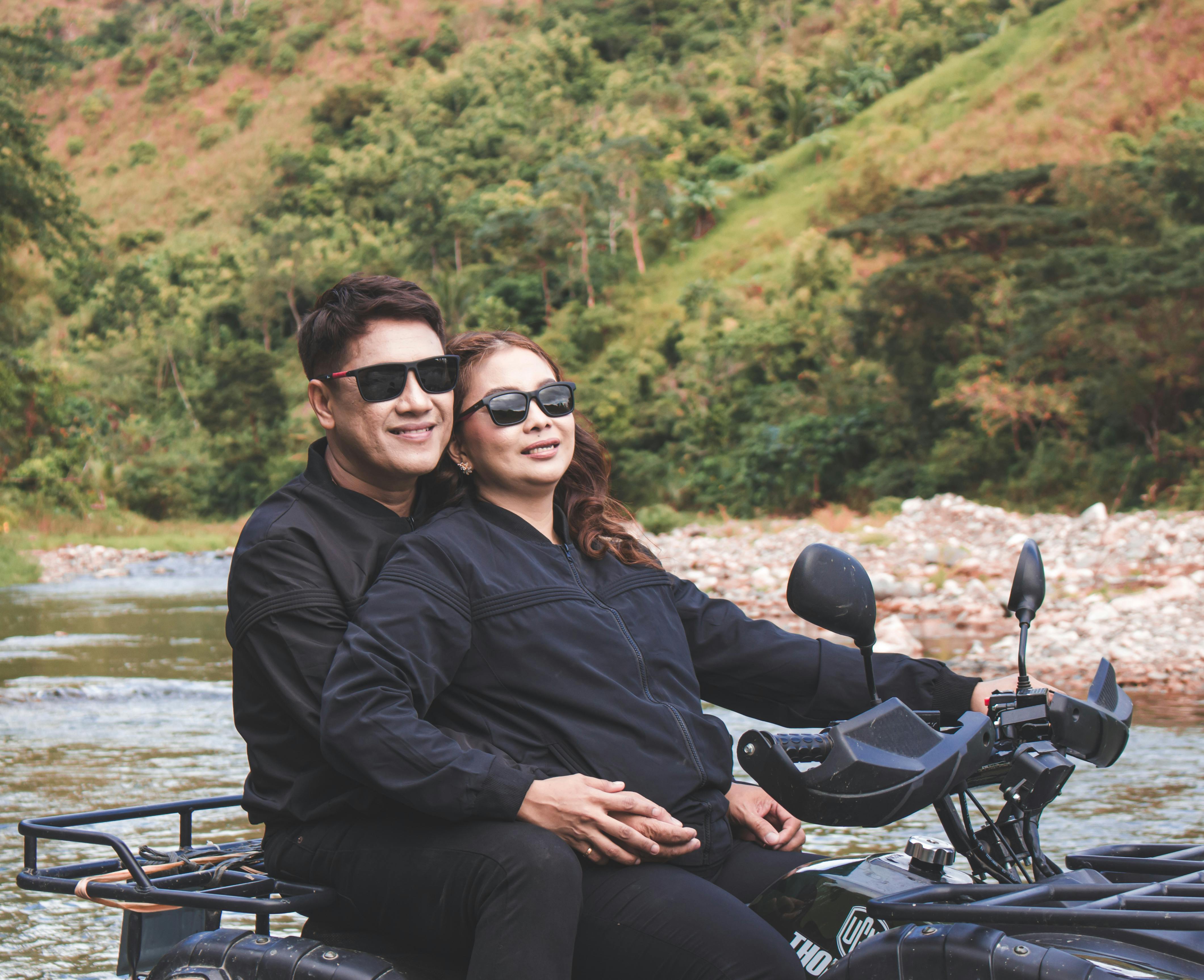 Couple riding an ATV by a stream in a lush valley, enjoying a scenic outdoor adventure.