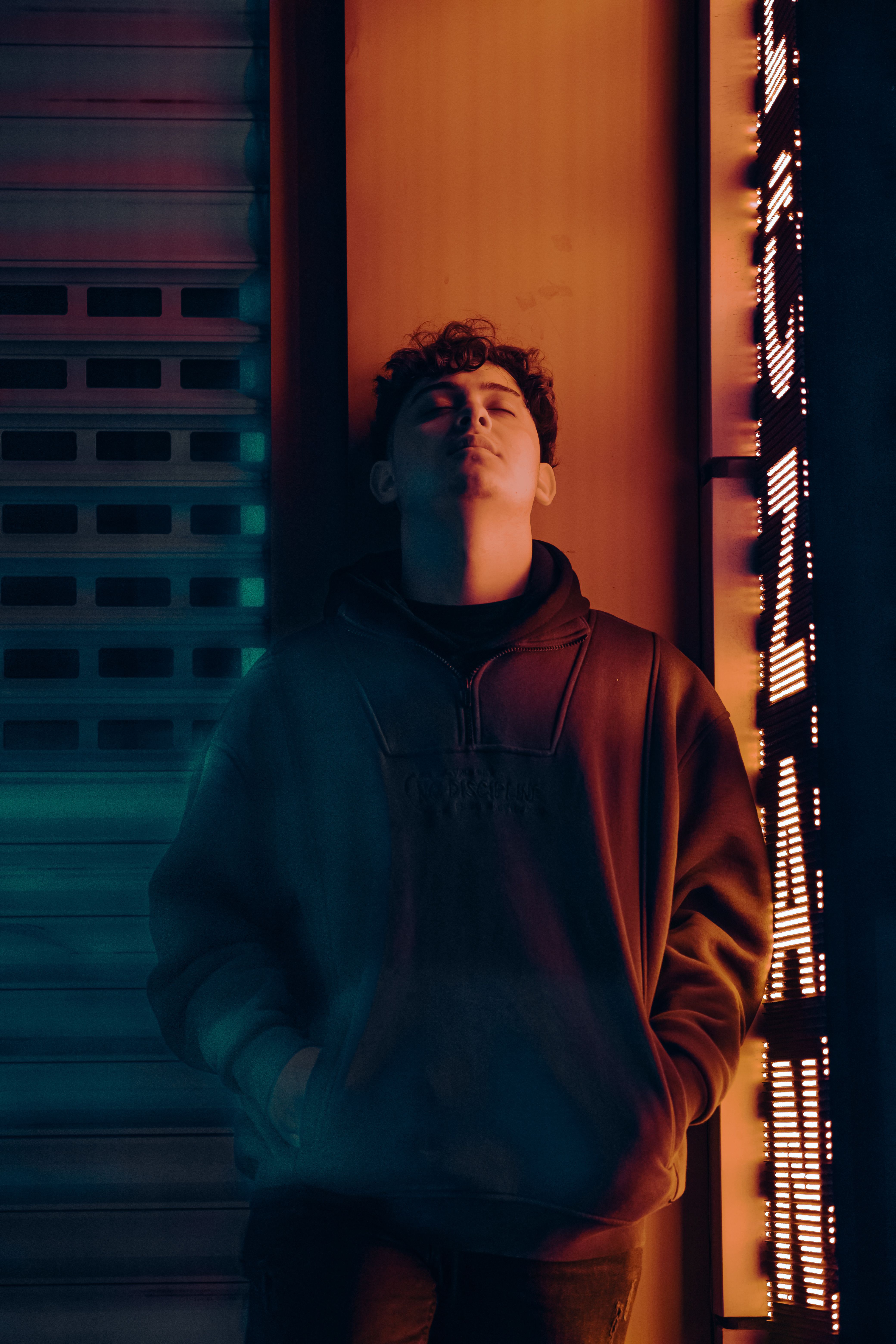 Young man with curly hair stands pensively with eyes closed by a neon-lit urban backdrop.