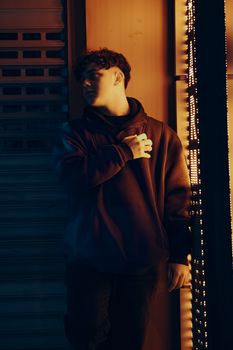 A young man in a black hoodie poses thoughtfully under neon lights.