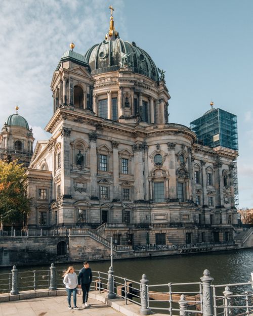Free A couple strolls along the riverbank by Berlin Cathedral, under a clear sky. Stock Photo