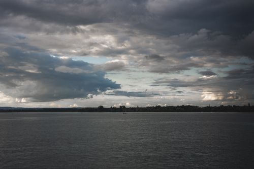 Free A tranquil lake scene with a lone sailboat under a dramatic cloudy sky, showcasing nature's beauty. Stock Photo