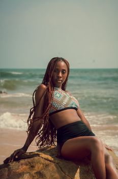A stylish woman with braided hair sits on a rock by the beach, exuding summer vibes.