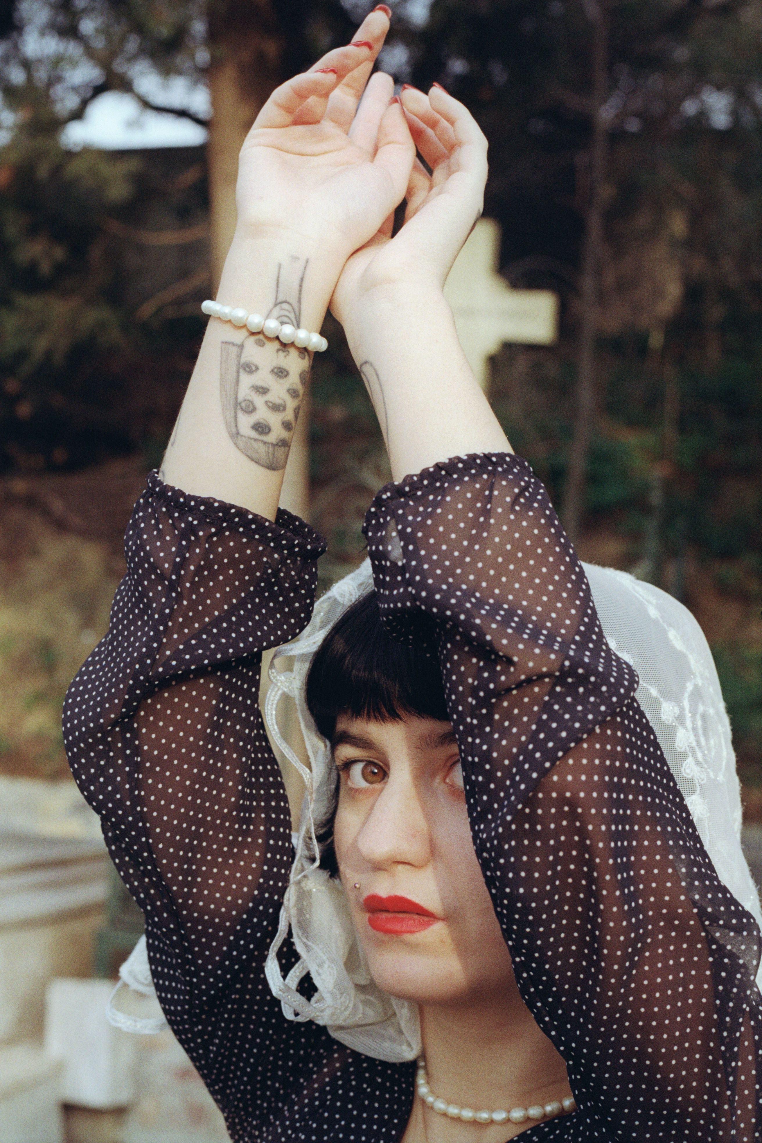 A woman poses outdoors with tattoos, veil, and raised arms, exuding an artistic and contemplative mood.
