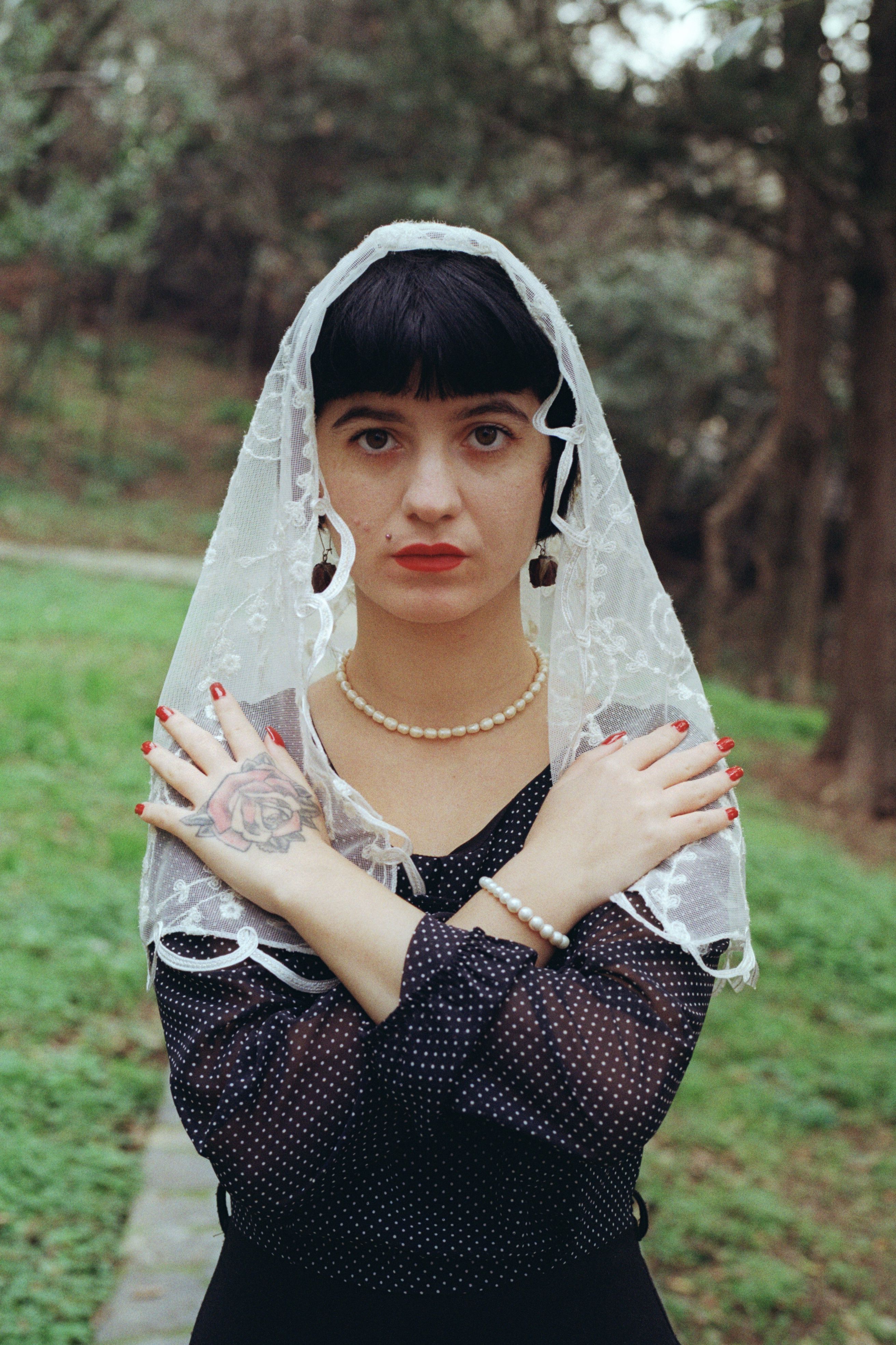 Portrait of a woman with a lace veil and tattoos, standing amidst greenery.