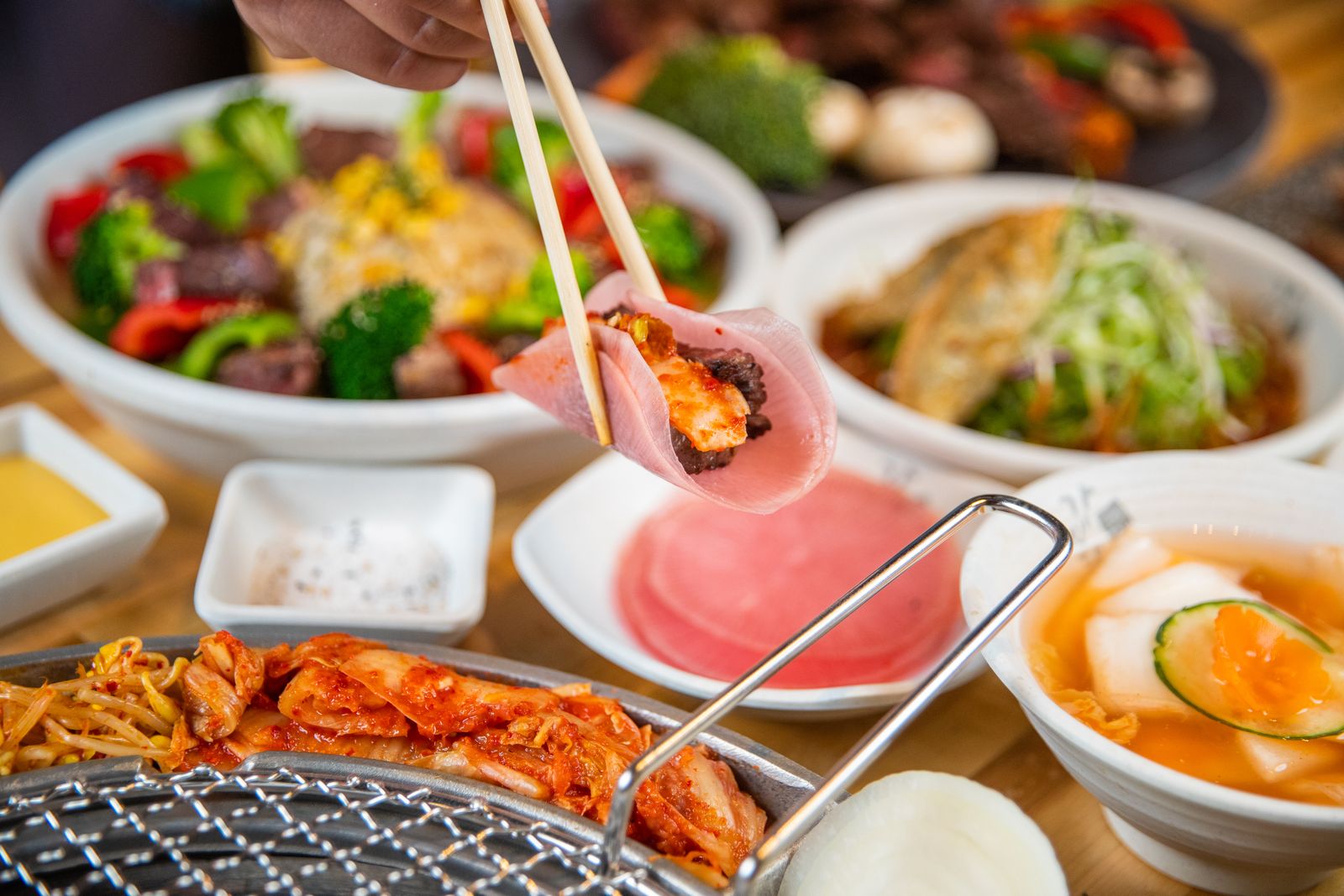 A Korean BBQ table spread — chopsticks lifting grilled meat over kimchi, banchan side dishes, dipping sauces, and a bowl of stew.