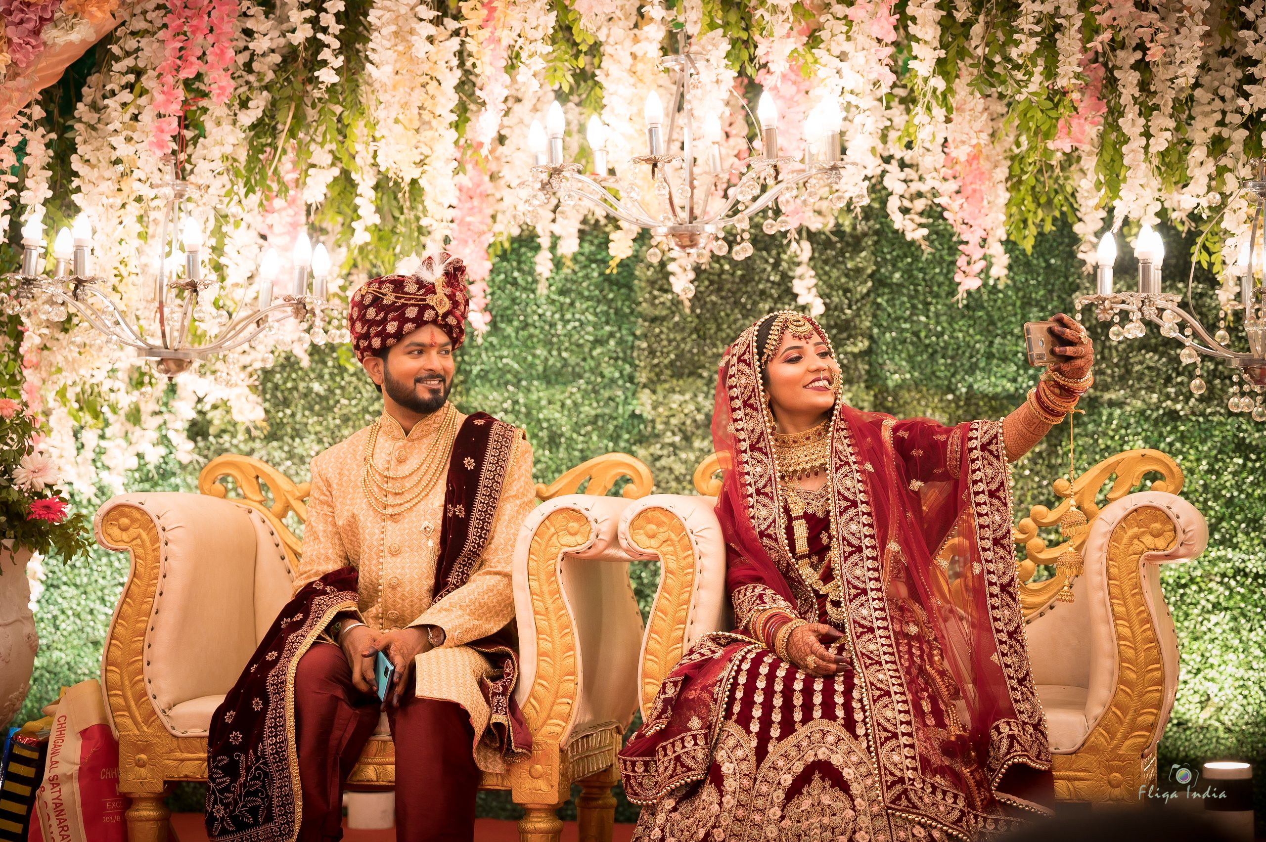 A couple in traditional wedding attire joyfully takes a selfie while seated in a beautifully decorated setting.