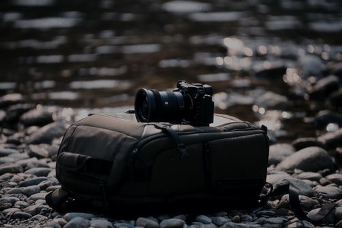 Free A DSLR camera rests on a backpack beside a tranquil pebbled river, captured in natural light. Stock Photo