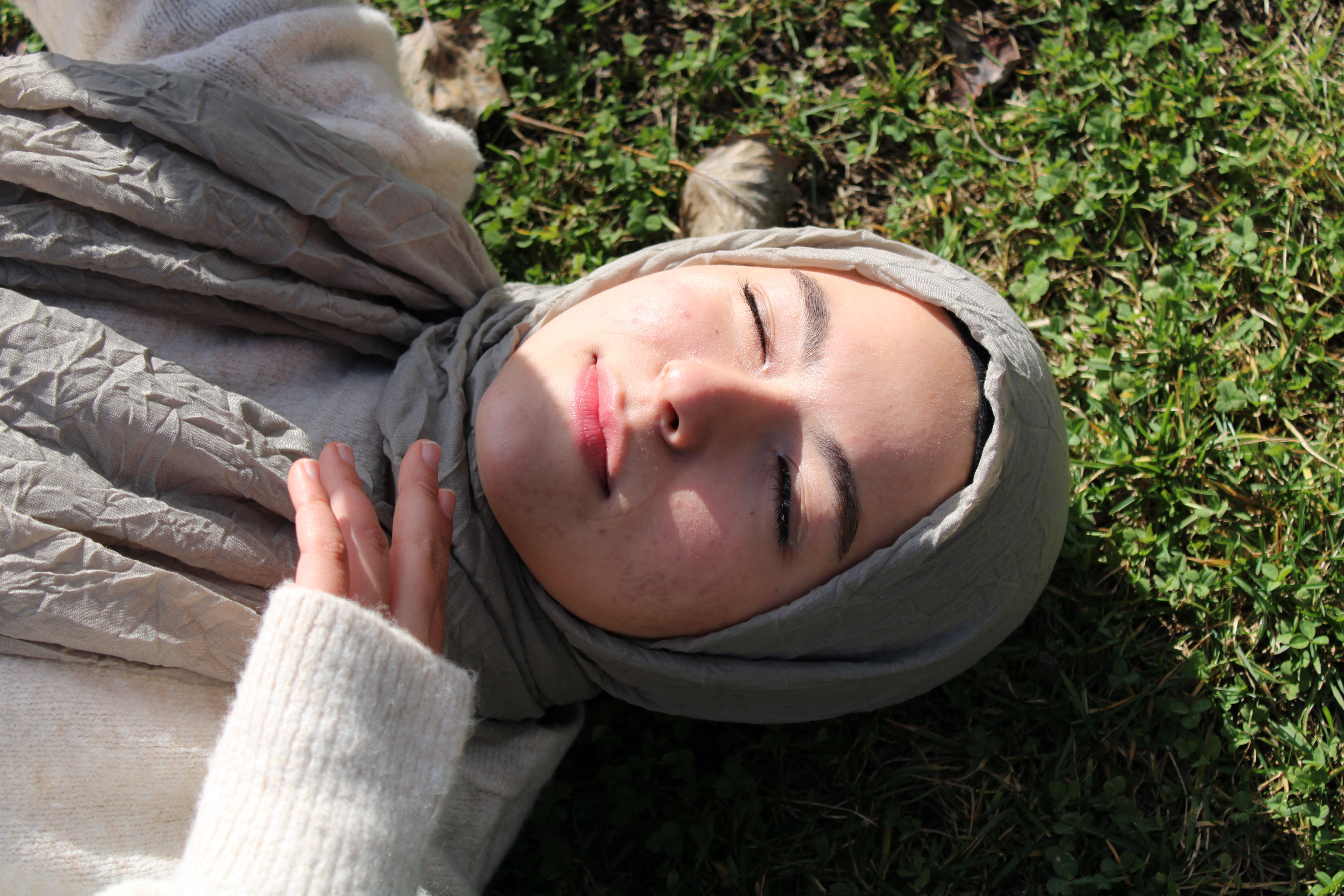 Asian woman in a hijab lying on grass, eyes closed in peaceful relaxation, photographed from above.