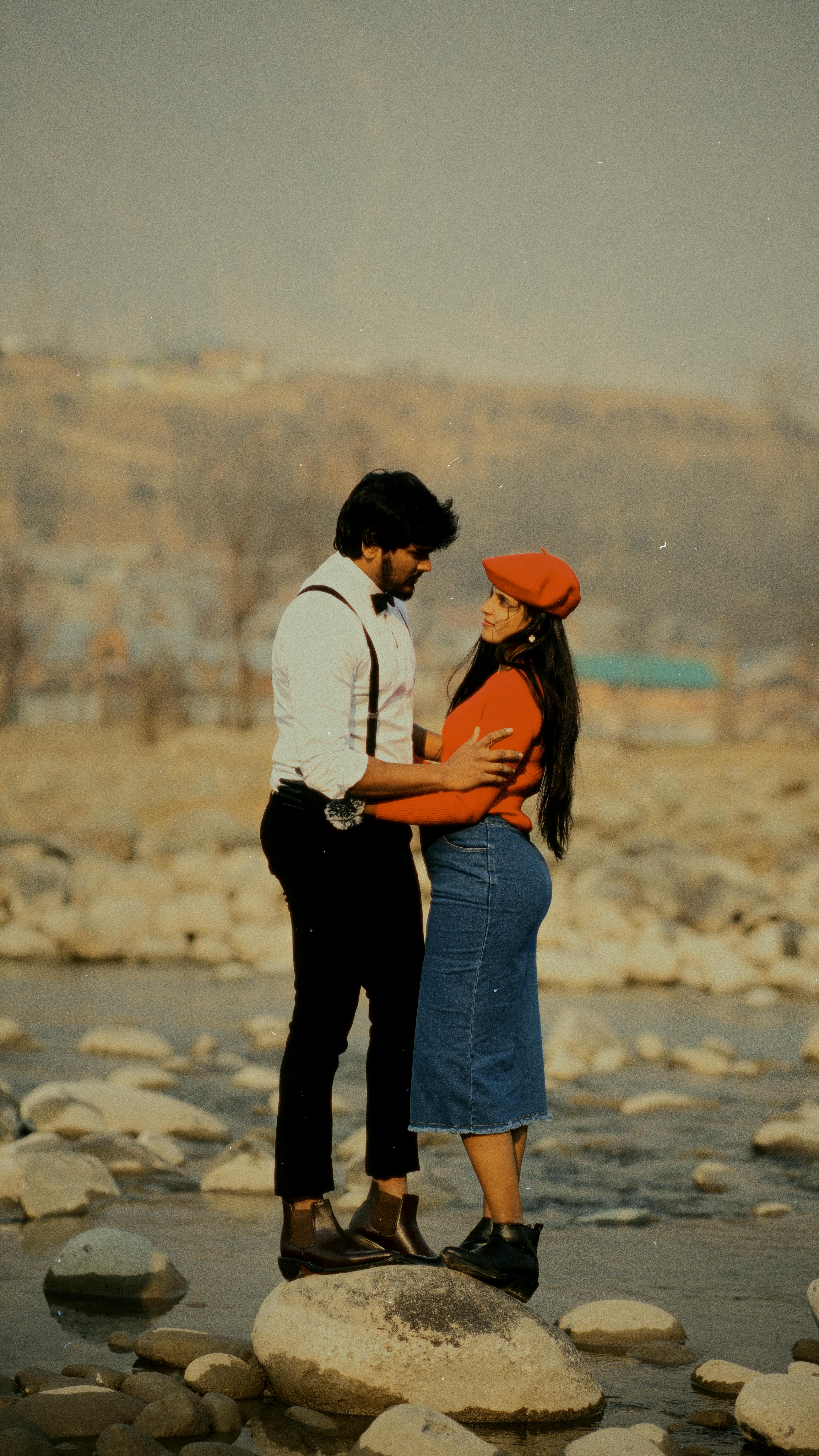 A loving couple embraces on a rocky riverbed, surrounded by a serene landscape.