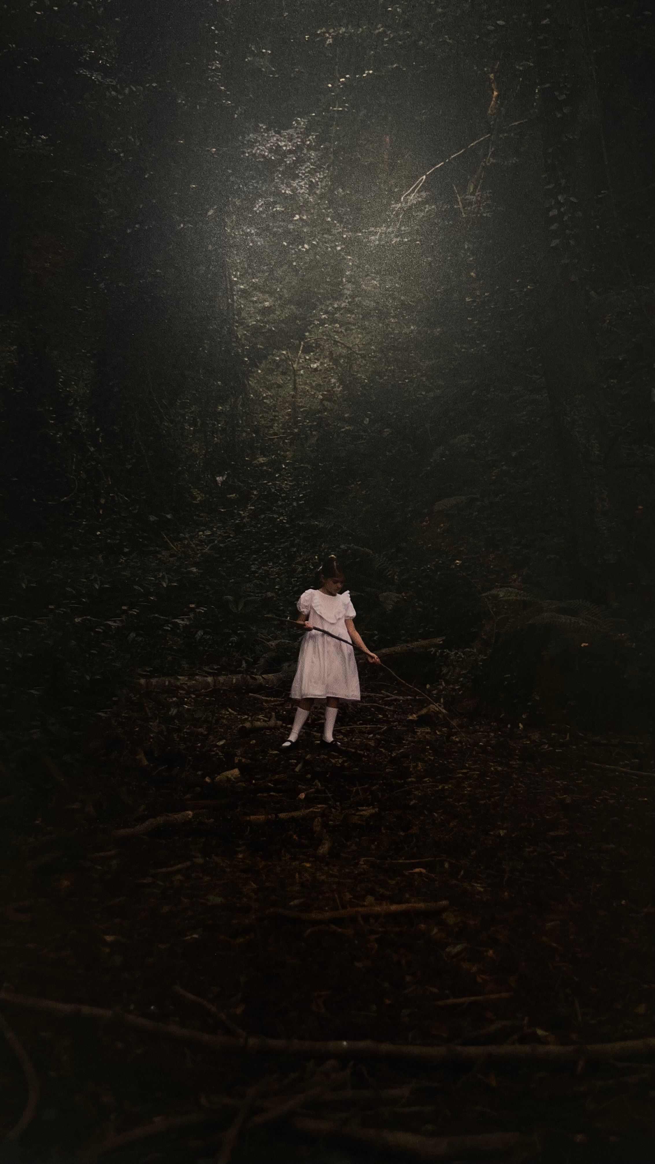 A young girl in a white dress standing alone in a dark forest. Eerie atmosphere.